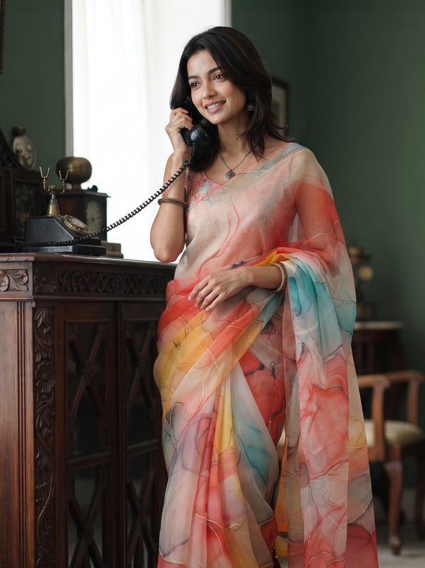 A woman wearing a lightweight multicolor printed saree stands indoors near a dark wooden cabinet, holding a black rotary telephone. The saree has an abstract watercolor-like pattern in soft pink, orange, yellow, blue, and white tones.