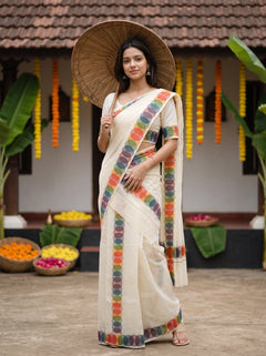A woman wearing a cream-colored cotton saree with colorful circular patterned borders and holding a large woven straw hat, standing in front of a traditional house decorated with flower garlands.