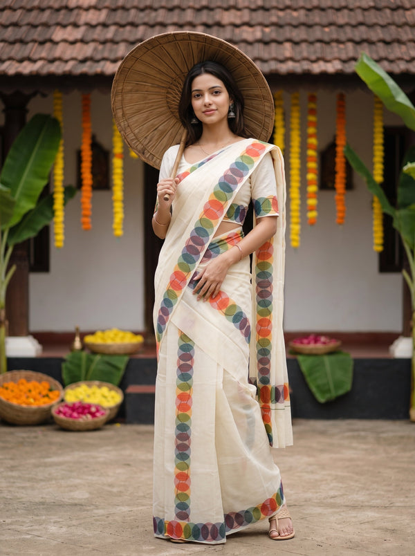 A woman wearing a cream-colored cotton saree with colorful circular patterned borders and holding a large woven straw hat, standing in front of a traditional house decorated with flower garlands.