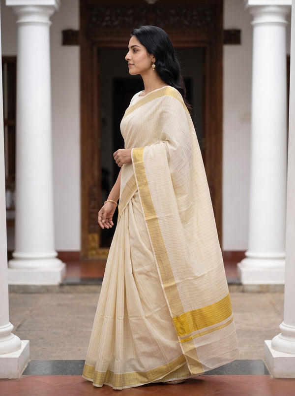 A woman wearing a light beige saree with a yellow-gold border, standing in a corridor with white columns.