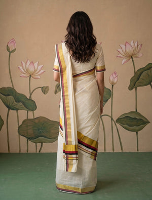 A woman viewed from behind wearing a cream-colored cotton saree with yellow, red, and purple striped borders, standing in front of a beige wall decorated with lotus motifs.