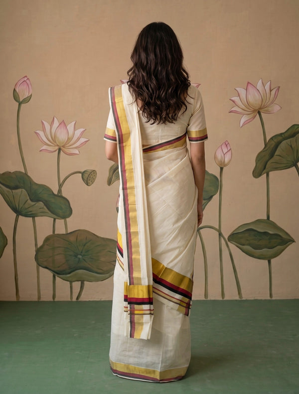A woman viewed from behind wearing a cream-colored cotton saree with yellow, red, and purple striped borders, standing in front of a beige wall decorated with lotus motifs.