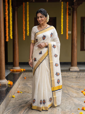 A woman wearing a white cotton saree with gold and blue printed motifs, standing in a traditional courtyard decorated with hanging marigold garlands.