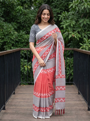 A woman wearing a coral pink and gray patterned saree with a matching gray printed blouse, standing on a wooden deck with green foliage in the background.