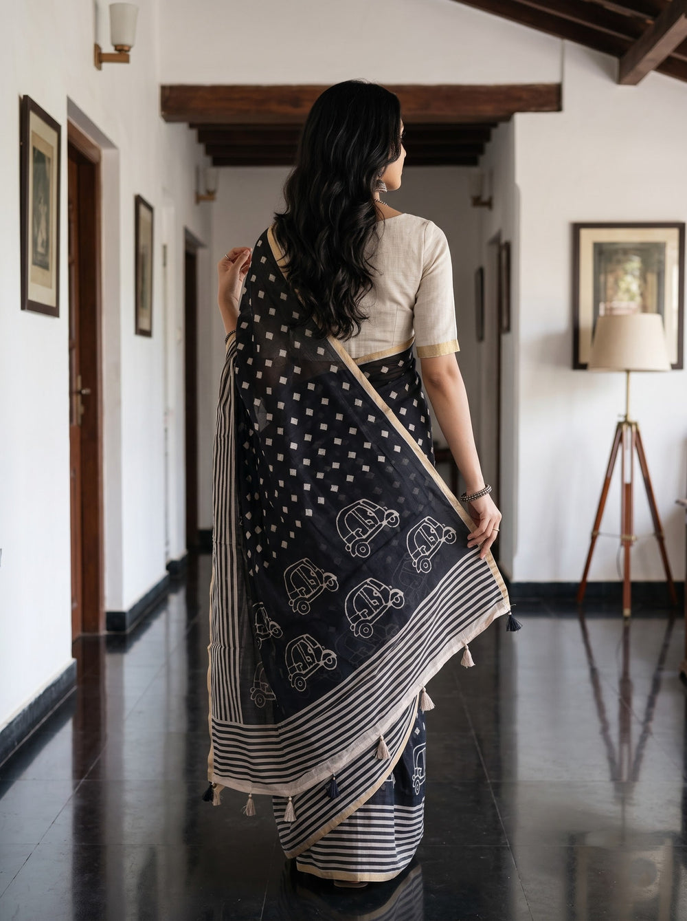 A woman modeling a black and white printed saree with a white blouse, standing in a traditional indoor hallway with wooden pillars and furniture.