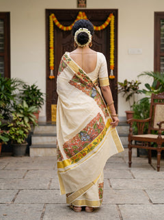 A woman viewed from behind wearing a cream-colored saree with a colorful floral border and a matching cream blouse, standing in front of a decorated doorway with plants and a chair in the background.