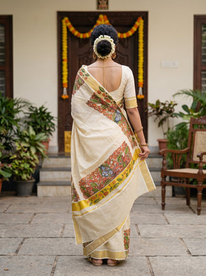 A woman viewed from behind wearing a cream-colored saree with a colorful floral border and a matching cream blouse, standing in front of a decorated doorway with plants and a chair in the background.