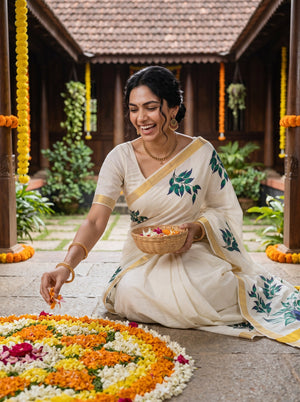 A woman in a cream-colored sari with green floral embroidery sits on a stone courtyard floor, placing flower petals into a decorative rangoli design. The background features traditional wooden structures and hanging marigold garlands.