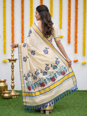 A woman wearing a cream-colored saree with blue and red floral patterns and a yellow border, standing in front of a decorated wall with marigold garlands and a brass lamp.