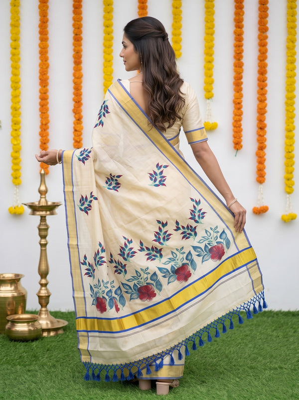 A woman wearing a cream-colored saree with blue and red floral patterns and a yellow border, standing in front of a decorated wall with marigold garlands and a brass lamp.