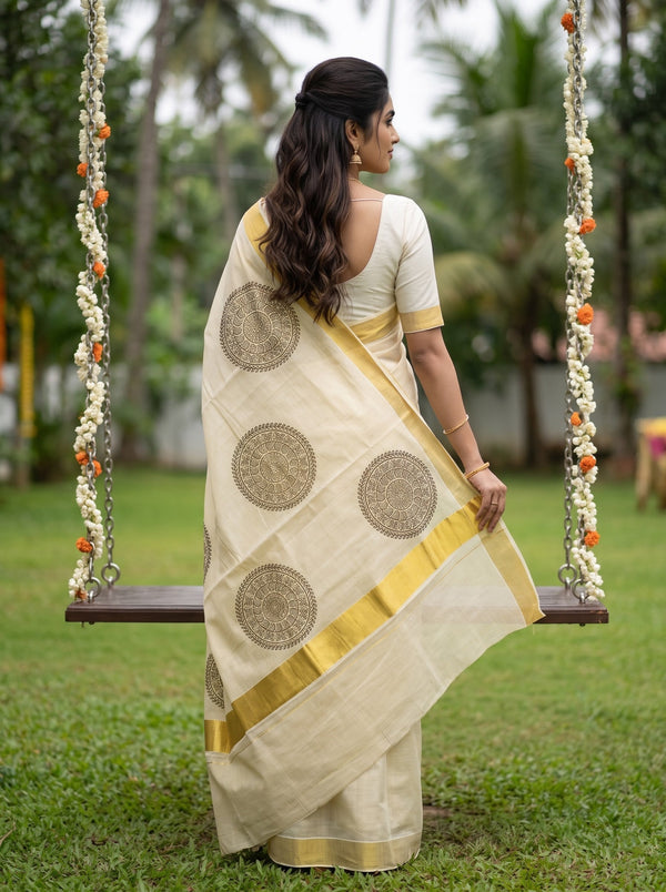 A woman stands on a swing in a garden wearing a cream-colored saree with gold border and circular motifs, viewed from behind.