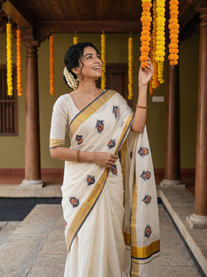 A woman wearing a cream-colored sari with gold borders and small floral motifs, standing in a traditional courtyard with hanging marigold garlands.