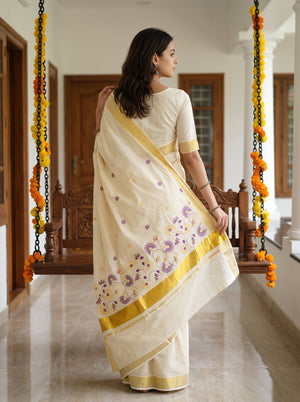 A woman wearing a cream-colored cotton saree with yellow borders and purple floral embroidery, standing in a decorated indoor hallway.