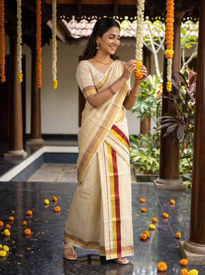 A woman wearing a cream-colored cotton saree with red and yellow stripes stands in a traditional courtyard, holding a garland.