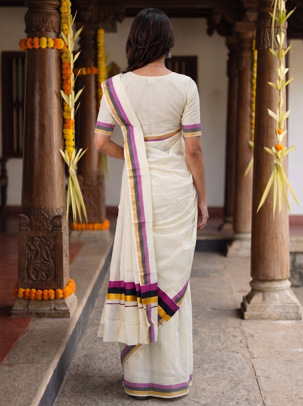 A woman viewed from behind wearing a cream-colored saree with purple, yellow, and orange border accents, standing in a traditional corridor with wooden pillars and garlands.