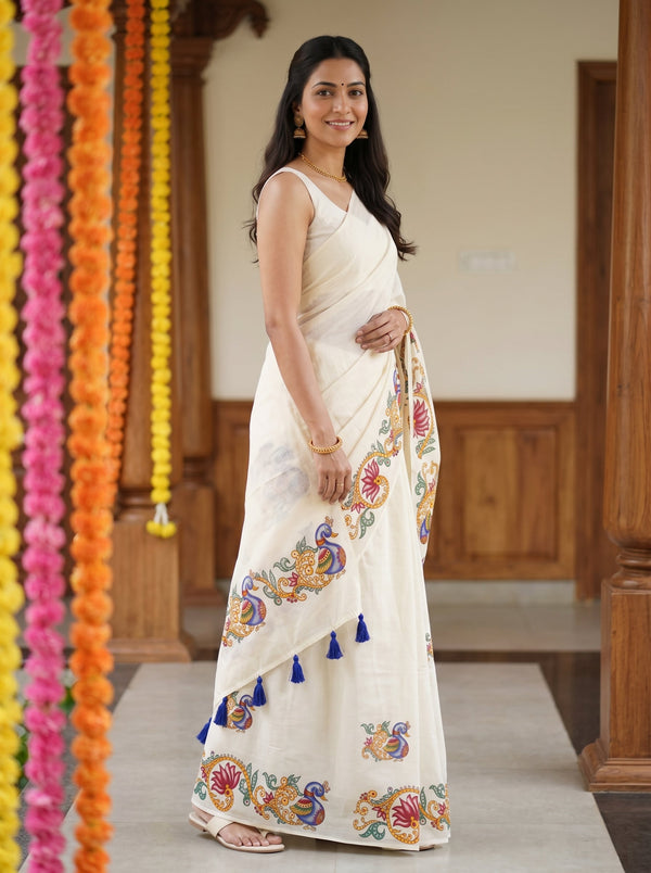 A woman wearing a cream-colored cotton saree with colorful floral embroidery and blue tassel accents, standing in a decorated indoor setting with garlands.