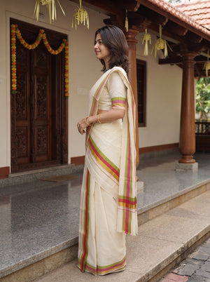 A woman wearing a cream-colored traditional Indian saree with green and red stripes, standing on stone steps in front of a house with wooden doors and hanging decorations.
