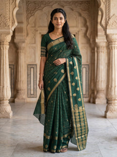 A woman wearing a dark green silk saree with gold zari patterns and a matching green blouse, standing in front of an ornate beige stone archway.