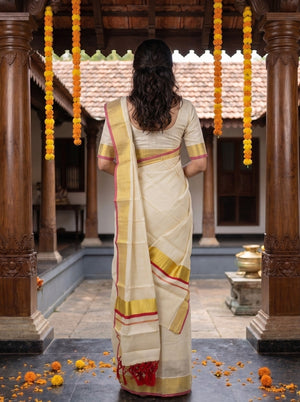 A woman viewed from behind wearing a cream-colored traditional saree with gold and red border details, standing in a courtyard with wooden pillars and hanging orange marigold garlands.