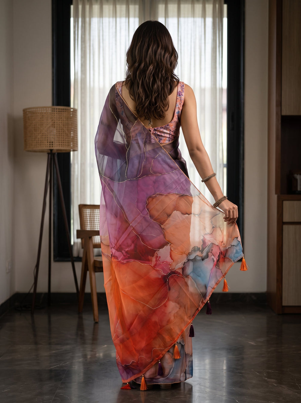 A woman wearing a colorful abstract-print saree in shades of orange, pink, purple, and blue, standing in a modern indoor room with large windows and curtains.