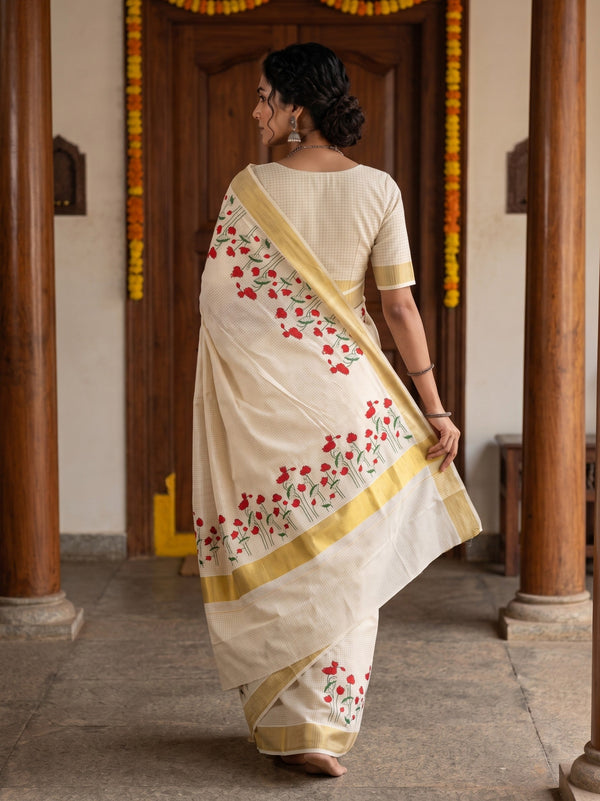 A woman wearing a cream-colored cotton saree with red floral embroidery and gold borders, shown from behind in a traditional indoor setting with wooden pillars.