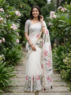 A woman wearing a white printed saree with pink and gray floral patterns stands on a stone path surrounded by green garden plants and pink flowers.