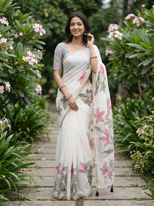 A woman wearing a white printed saree with pink and gray floral patterns stands on a stone path surrounded by green garden plants and pink flowers.
