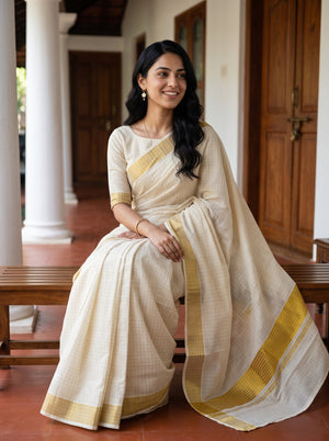 A woman wearing a cream-colored cotton saree with a yellow-gold border sits on a wooden bench in a traditional house corridor.