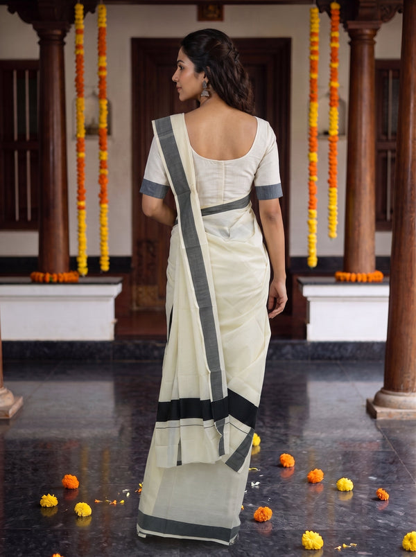 A woman wearing a cream-colored cotton saree with dark gray borders and a matching cream blouse, standing in a traditional courtyard decorated with marigold garlands.