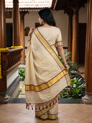 A woman wearing a cream-colored saree with gold and maroon borders, standing in a traditional courtyard with wooden pillars.