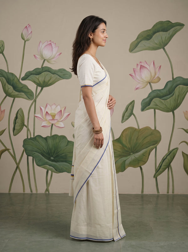 A woman wearing a cream-colored cotton saree with thin blue and gold borders, standing in profile against a wall decorated with a large-scale green lotus leaf and pink lotus flower mural.