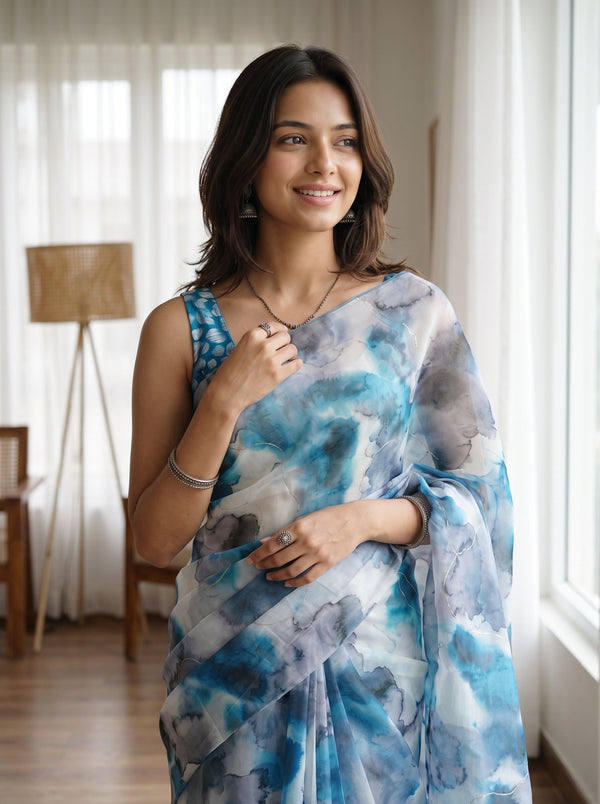 A woman wearing a blue and white watercolor-patterned saree with a matching blue patterned sleeveless blouse, standing in a brightly lit room with sheer curtains and a floor lamp.