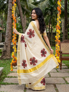 A woman wearing a cream-colored cotton saree with yellow borders and dark red floral prints, standing on a stone path in a garden next to a swing decorated with yellow marigold garlands.