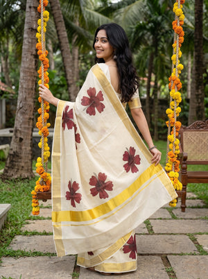 A woman wearing a cream-colored cotton saree with yellow borders and dark red floral prints, standing on a stone path in a garden next to a swing decorated with yellow marigold garlands.