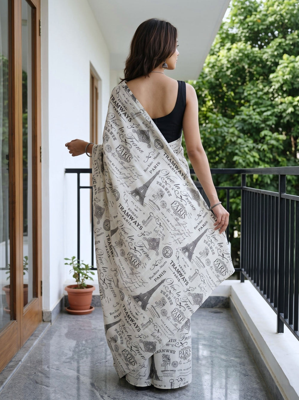 A woman modeling a newspaper-print saree with a black sleeveless blouse, standing on a balcony with green trees in the background.