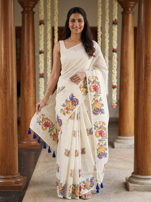 A woman wearing a cream-colored cotton saree with colorful floral and peacock motifs, standing in a corridor with wooden pillars.