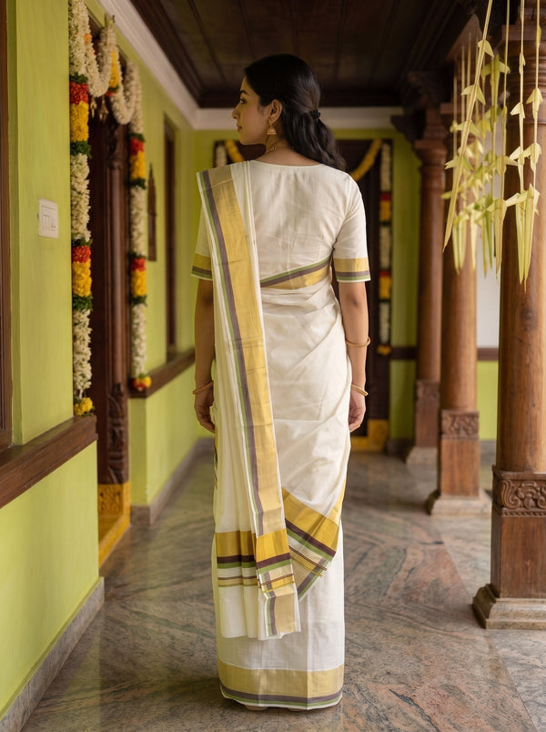 A woman viewed from behind wearing a white cotton saree with yellow, green, and gray border stripes, standing in a traditional corridor with wooden pillars and green walls.