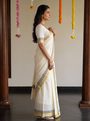 A woman wearing a traditional Indian saree in cream and gold, standing in profile in an indoor setting with decorative garlands hanging in the background.
