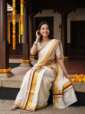 A woman wearing a cream-colored cotton saree with yellow, orange, and black stripes sits on stone steps in a traditional courtyard decorated with marigold garlands.