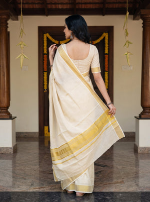 A woman wearing a cream-colored cotton saree with a yellow border and a matching cream blouse, photographed from behind in a traditional indoor setting with wooden pillars and garlands.