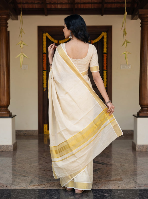 A woman wearing a cream-colored cotton saree with a yellow border and a matching cream blouse, photographed from behind in a traditional indoor setting with wooden pillars and garlands.