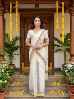 A woman stands on stone steps in front of a carved wooden doorway, wearing a cream-colored sari with blue and gold patterned borders. Yellow flower garlands hang from the doorway, and potted plants and scattered yellow petals decorate the surroundings.