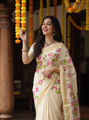 A woman wearing a cream-colored floral saree with pink and green printed motifs, standing in a decorated indoor setting with yellow and orange marigold garlands hanging from above.