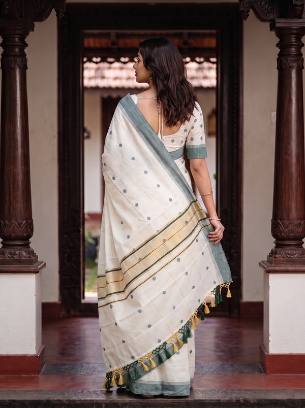 A woman wearing a white cotton saree with sage green border and gold stripes, standing in a traditional courtyard framed by dark wooden pillars.