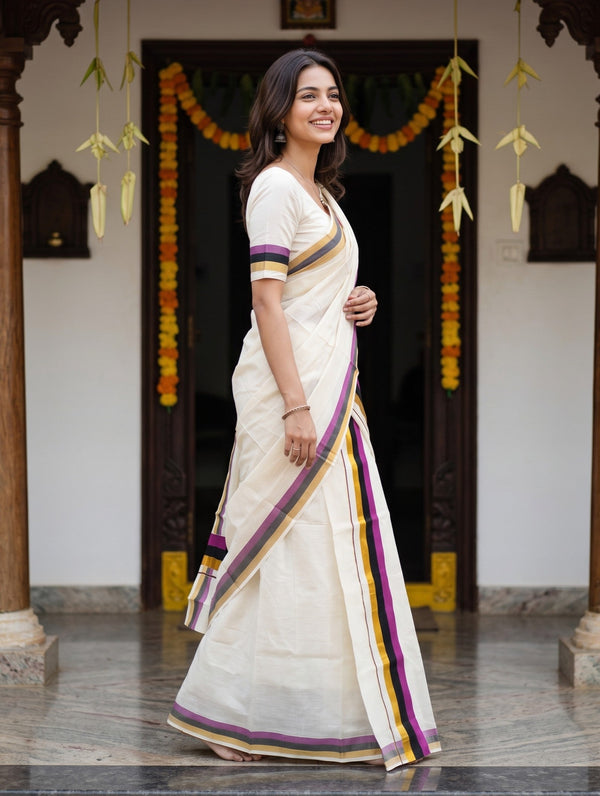 A woman wearing a white cotton saree with purple, yellow, and gold borders, standing in a traditional courtyard with wooden pillars and garlands.