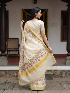 A woman wearing a cream-colored saree with pink floral prints and gold borders, standing in front of a traditional building doorway.