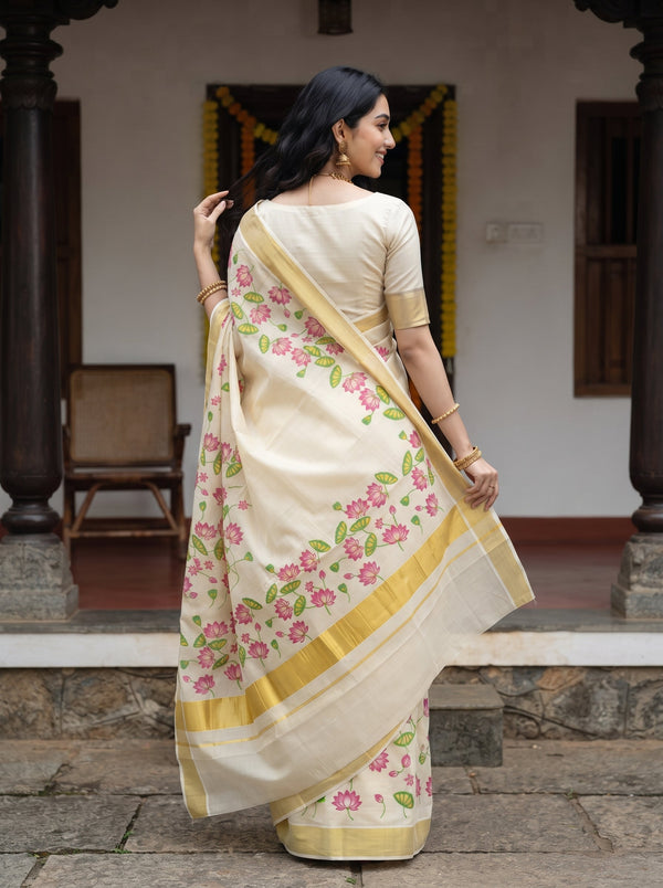 A woman wearing a cream-colored saree with pink floral prints and gold borders, standing in front of a traditional building doorway.