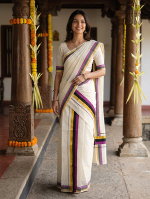 A woman wearing a cream-colored sari with purple, yellow, and green striped borders stands in a traditional corridor with wooden pillars decorated with marigold garlands.