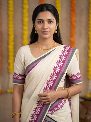 A woman wearing a cream-colored sari with purple and gray floral border patterns, paired with a matching cream blouse and gold jewelry, set against a warm background with hanging marigold garlands.