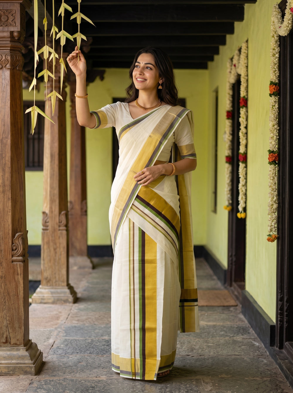 A woman wearing a traditional Indian saree stands in a corridor with wooden pillars. The saree is off-white/cream with a mustard yellow border and green stripes, paired with a matching off-white blouse.